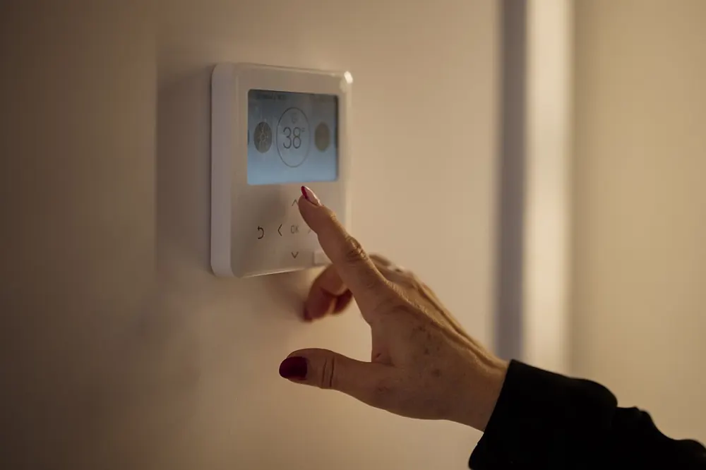 A hand adjusting a wall mounted digital thermostat in a dimly lit room